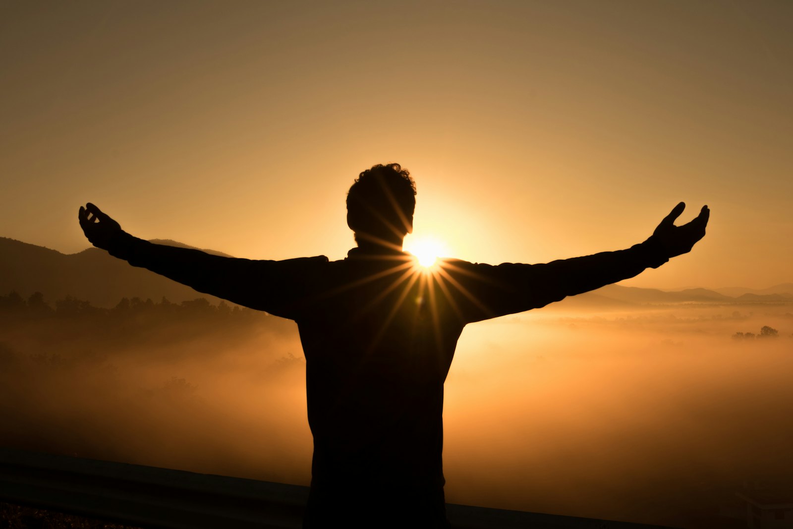 A person standing with arms wide open as the sun rises over mist-covered hills.